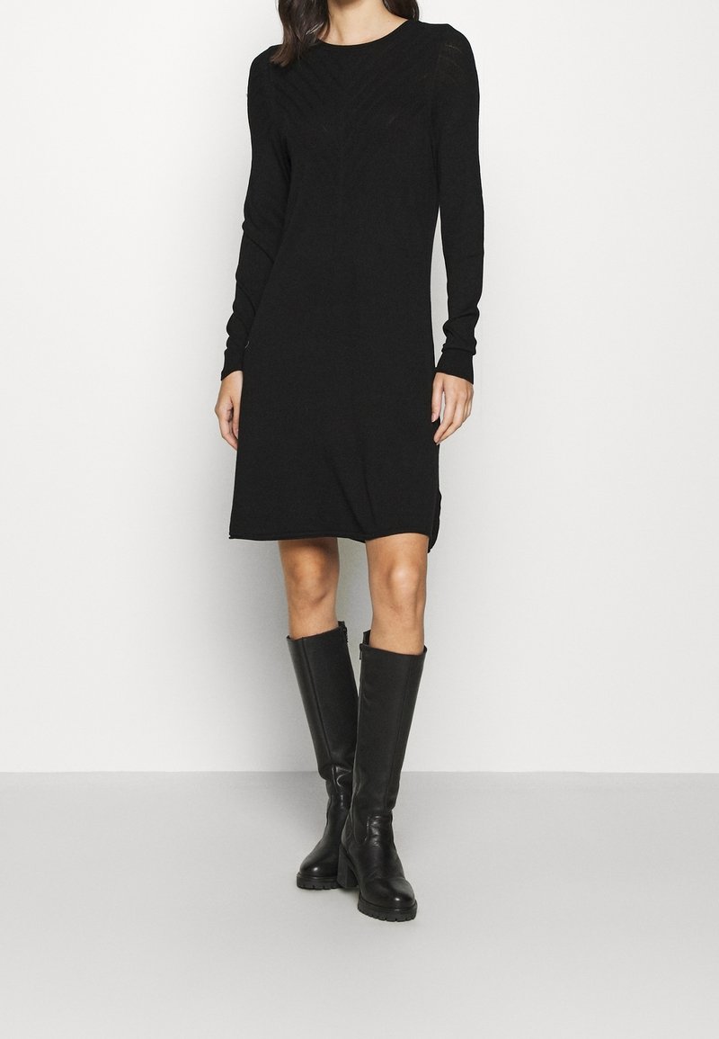 Schwarzes Langarmkleid mit einer figurbetonten Silhouette, geripptem Textur an den Schultern und einem gewellten Saum. Kombiniert mit knielangen schwarzen Stiefeln.