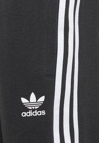 Juodos sportinės kelnės su trimis baltomis juostelėmis šonuose, turinčios baltą Adidas logotipą ir lygią tekstūrą.