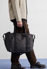 Sac de sport noir imperméable avec poignées doubles et bandoulière. Présente un corps texturé et des surpiqûres contrastées sur le bord inférieur.