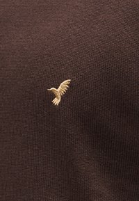 Pull en maille marron foncé avec un petit logo oiseau beige brodé sur le côté gauche. Texture lisse avec un motif subtil et uniforme.