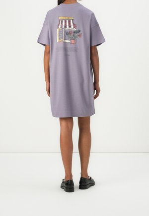 Persona que lleva un vestido suelto de manga corta color lavanda con un gráfico colorido de una tienda y una maceta en la espalda, combinado con zapatos negros.