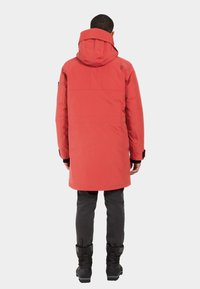 Röd vadderad jacka med huva, med rak design och justerbara ärmslut. Bärs över mörkgrå byxor och svarta stövlar.