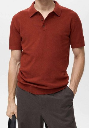 Mies, joka pukeutuneena ruosteenväriseen lyhythihaiseen polopaitaan, jossa on napit, tummat housut, musta laukku kädessä ja toinen käsi taskussa.