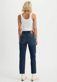 Jeans in denim blu a vita alta con taglio dritto, dotati di tasche posteriori e un'etichetta in pelle del marchio. Indossati con un top bianco senza maniche.