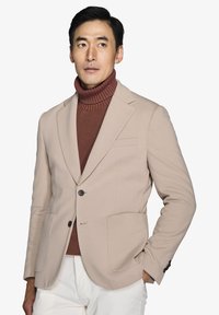 Beige blazer med to store frontlommer, enkelt knaplukning, notched lapels og en blød tekstur, lagd oven på en rustfarvet rullekrave sweater.