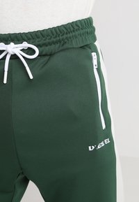 Gröna joggingbyxor med en vit sidpanel, elastisk midja, dragsko och fickor med dragkedja. Har en logotyp tryckt på benet.