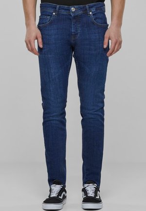 Jeans de mezclilla azul oscuro con un ajuste slim, que presentan bolsillos frontales, cierre de cremallera y un botón de metal en la cintura, combinados con zapatillas negras.