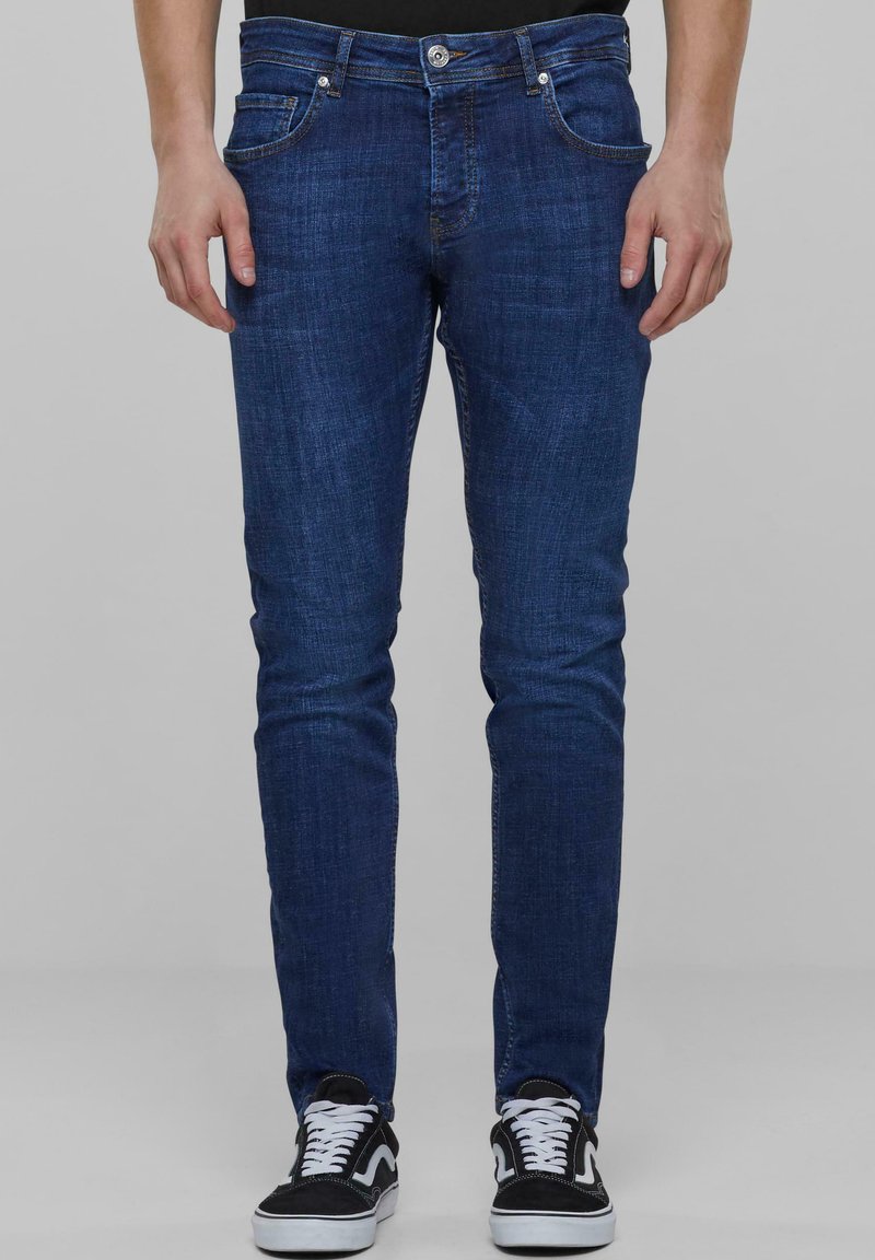 Tumed sinised denim püksid kitsaste lõigetega, esi-taskutega, luku ja metallnööpiga vööl, koos mustade tossudega.