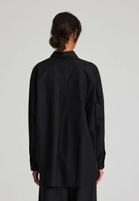 Maglietta nera a maniche lunghe con spalla posteriore, patta con bottoni frontale e vestibilità comoda. Realizzata in un tessuto liscio e leggero.