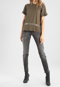 Camiseta de algodón verde oliva con mangas cortas y un estampado gráfico blanco en el dobladillo. Combinada con vaqueros ajustados grises y botas negras hasta el tobillo.