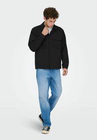 Homme portant une veste noire à boutons, un jean bleu clair et des baskets noires, regardant vers le bas en marchant sur un fond blanc uni.