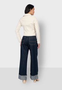 Witte cropped jacket met lange mouwen, gecombineerd met donkerblauwe brede jeans met opgerolde pijpen en achterzakken, bekeken vanuit de achterkant.