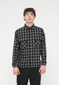 Πουκάμισο από φανέλα με ασπρόμαυρο καρό σχέδιο, δύο τσέπες στο στήθος, κουμπώνει με κουμπιά μπροστά και έχει χρυσά κουμπιά. Απαλή υφή με αντιθετικές λεπτομέρειες στις τσέπες.