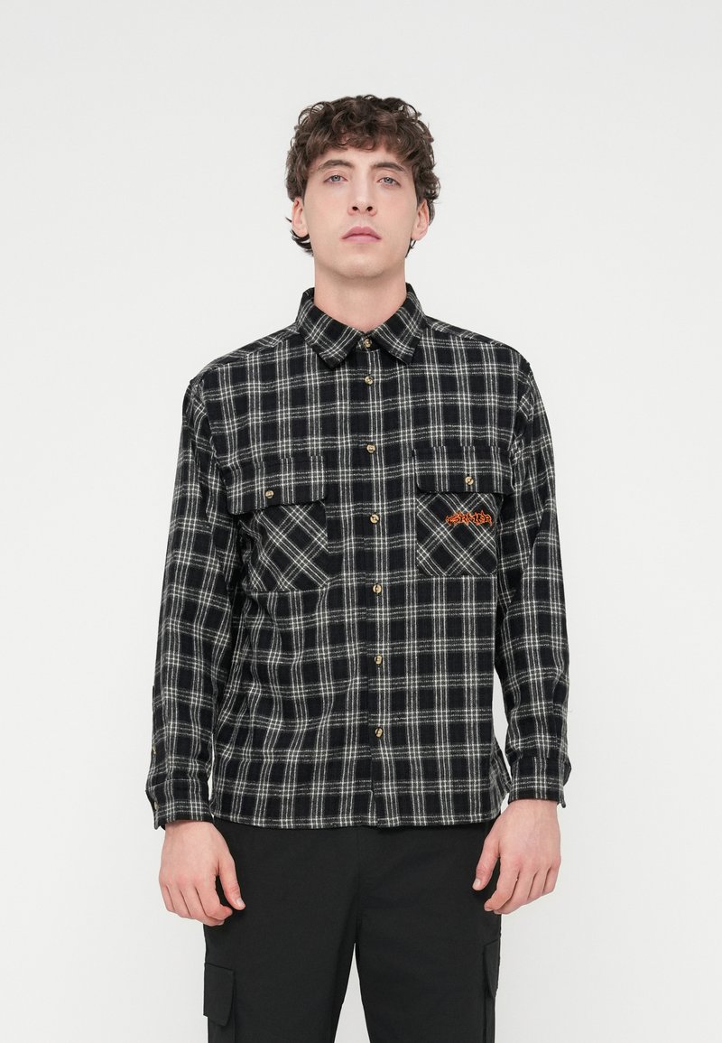 Πουκάμισο από φανέλα με ασπρόμαυρο καρό σχέδιο, δύο τσέπες στο στήθος, κουμπώνει με κουμπιά μπροστά και έχει χρυσά κουμπιά. Απαλή υφή με αντιθετικές λεπτομέρειες στις τσέπες.