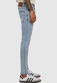 Light blue skinny jeans with a textured finish, five-pocket design, and a brown leather logo patch on the back waistband. Paired with white sneakers.