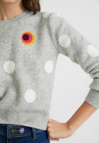 Pull en tricot gris avec des pois blancs, broderie circulaire colorée contrastante, et ourlet et poignets côtelés. Associé à un jean bleu.