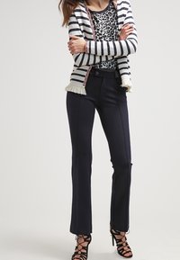 Cardigan a righe con frange, camicia a stampa leopardata, pantaloni fitted blu navy e tacchi neri con cinturini creano un look stratificato e testurizzato.