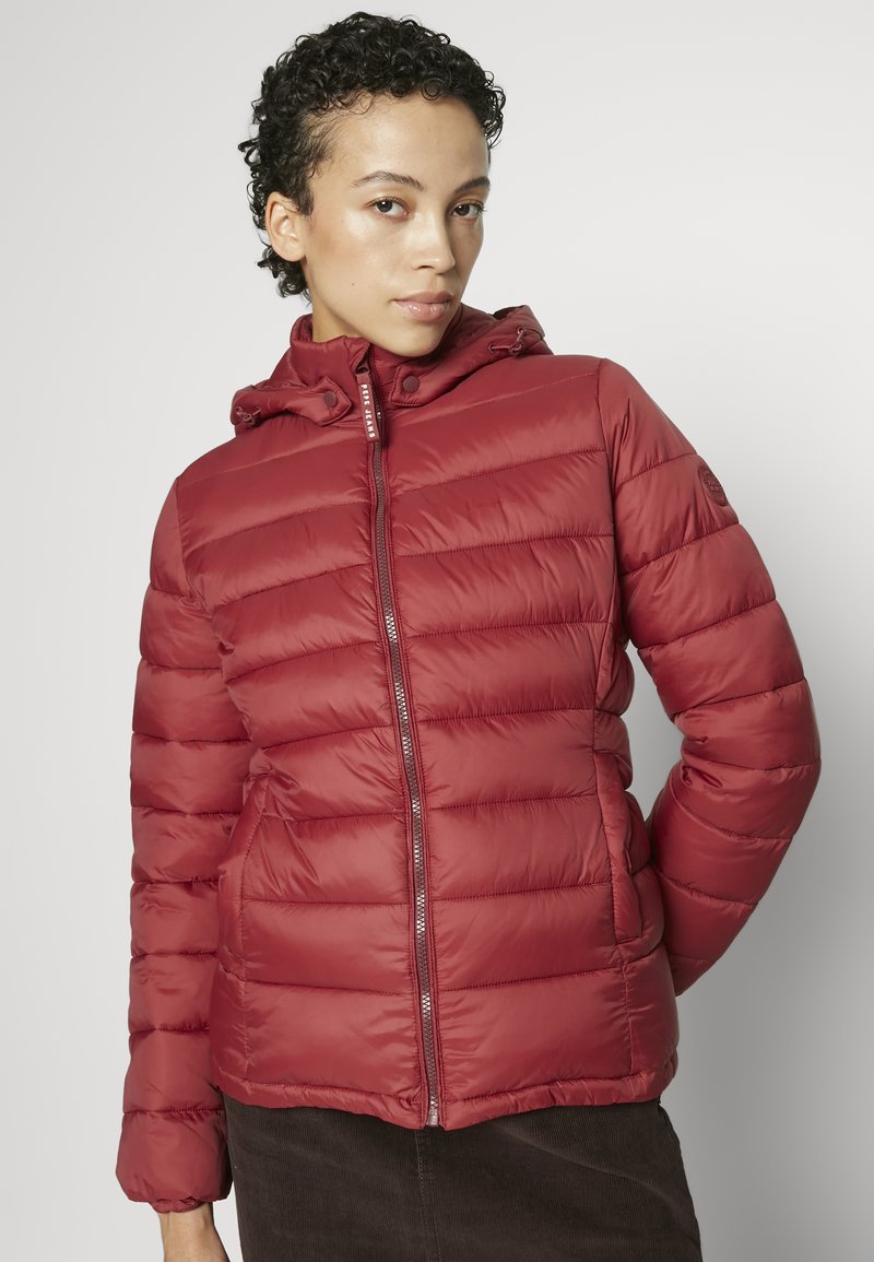 Mujer con cabello rizado, vestida con una chaqueta acolchada con capucha roja y una falda marrón, de pie contra un fondo ligero y sencillo.