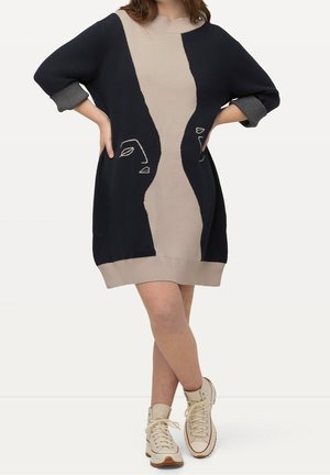 Femme portant une robe-pull longue noire et beige avec des motifs abstraits de visages, des manches grises retroussées et des baskets montantes crème, debout les jambes croisées.