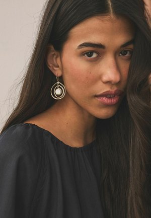 Young woman with long dark hair wearing black top and gold hoop earrings with white stone, looking thoughtfully toward the camera.