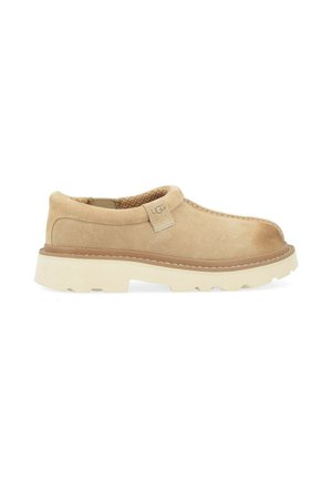 Chaussure beige en daim à enfiler avec semelle blanche épaisse à rainures et étiquette avec logo UGG près du col rembourré, vue de côté sur fond blanc.