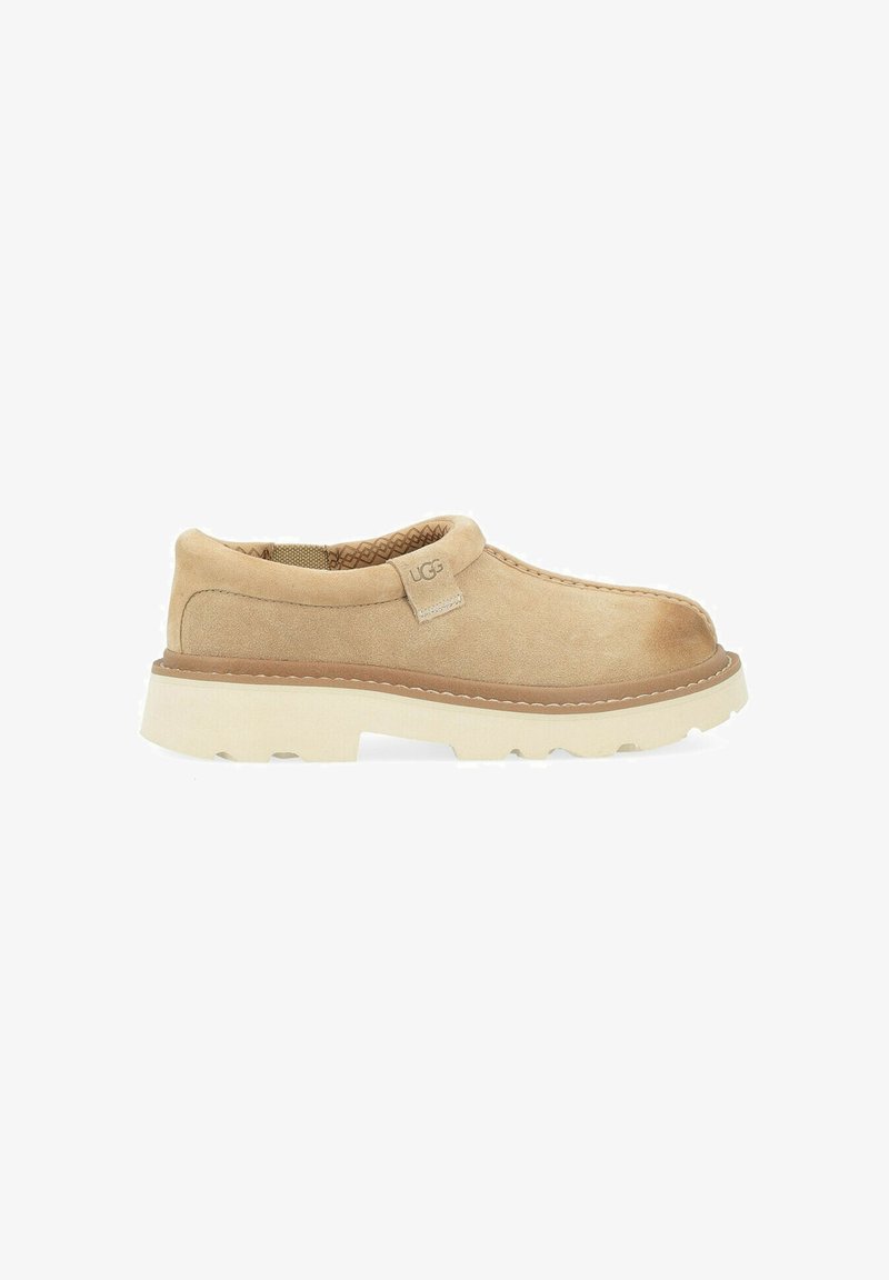 Chaussure beige en daim à enfiler avec semelle blanche épaisse à rainures et étiquette avec logo UGG près du col rembourré, vue de côté sur fond blanc.