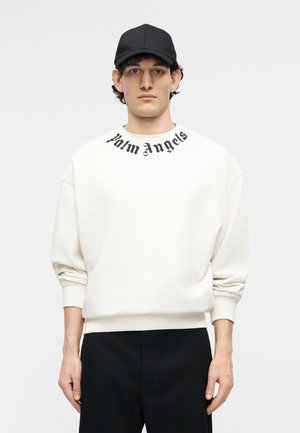 Άνδρας που φοράει λευκό φούτερ με το κείμενο «Palm Angels» κοντά στον γιακά, μαύρο παντελόνι και μαύρο καπέλο baseball, στέκεται μπροστά σε απλό φόντο.