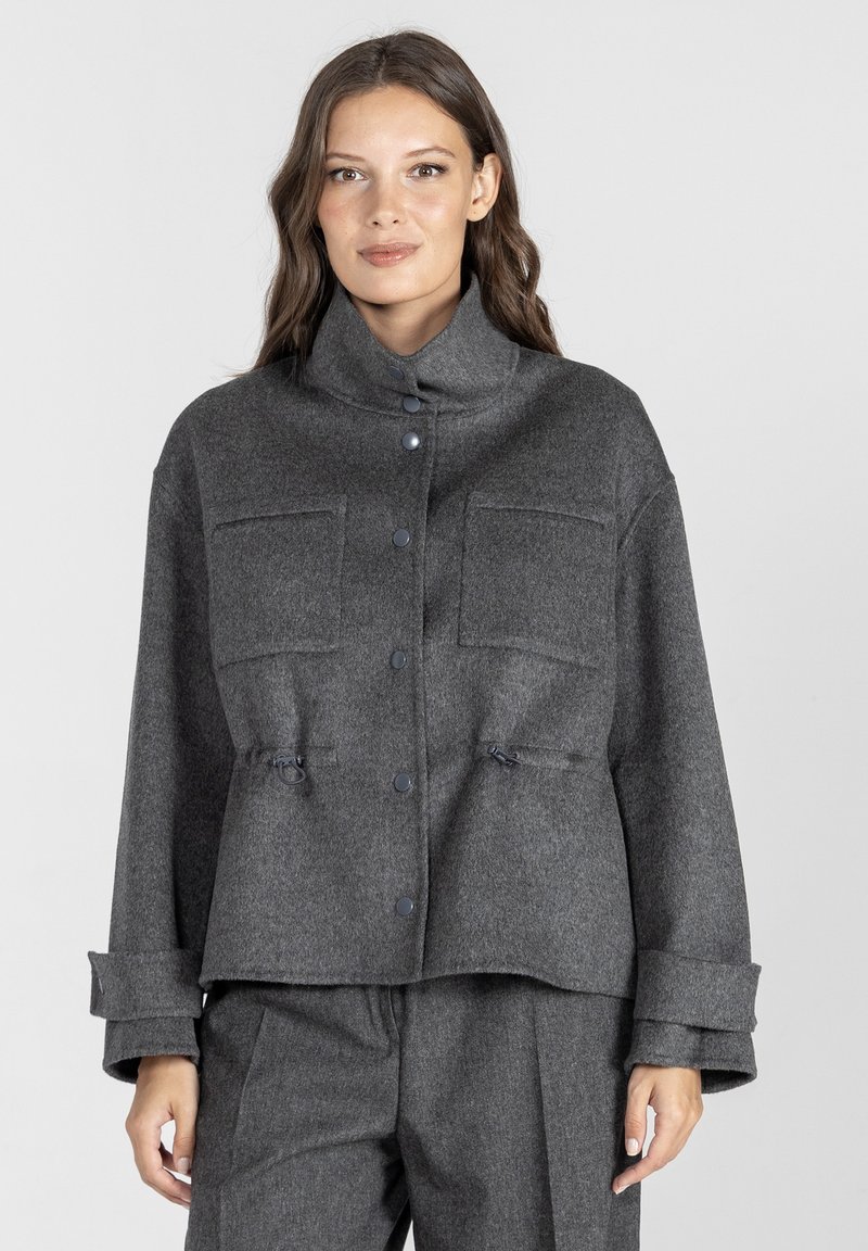 Szary wełniany żakiet z wysokim kołnierzem, guzikami z przodu, dwoma kieszeniami na piersiach oraz regulowanymi sznurkami w pasie. Teksturowane wykończenie.