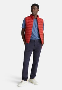 Muž stojící s rukama v kapsách, oblečený v červené prošívané vestě přes modré tričko, námořnických kalhotách a bílých teniskách.