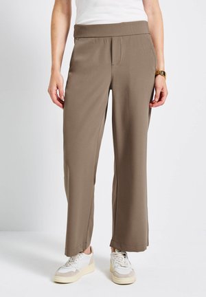 Bruine, wijde broek gemaakt van zacht materiaal met een elastische tailleband. Heeft voorzakken en een gladde texture. Gepaard met witte sneakers.
