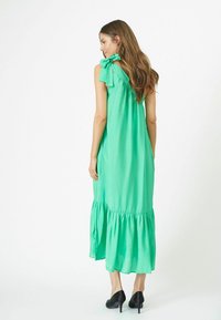 Rochie maxi verde cu detaliu de fundă pe umă, tiv clasic, textură fină și croială lejeră, asortată cu pantofi cu toc înalt negri.