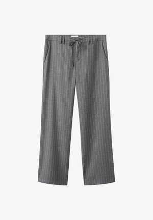 Mladá žena stojí s rukami v kapse, oblečená v voľnom sivom prúžkovanom obleku, čiernom krátkom topánke a hnedých topánkach na sivom pozadí.