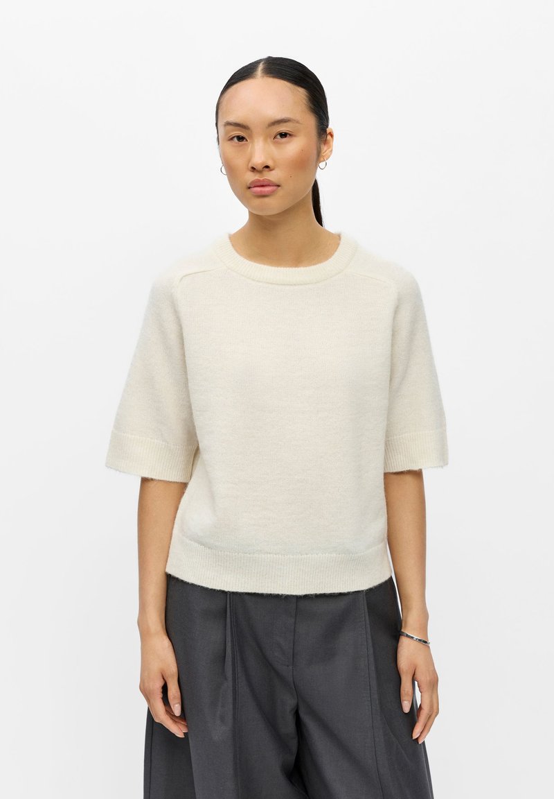 Jeune femme aux cheveux foncés attachés, portant un pull crème à manches courtes et un pantalon ample gris foncé sur fond blanc.