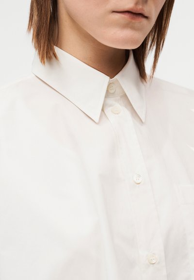 Λευκό πουκάμισο με κουμπιά, με μυτερό γιακά, μαλακή υφή και κλασικά κουμπώματα. Απλός σχεδιασμός με λεπτομέρεια τσέπης στο στήθος.