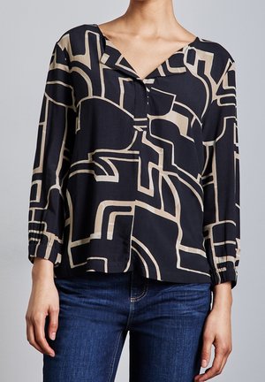 Kvinde iført en sort bluse med beige abstrakte geometriske mønstre og blå jeans, stående foran en ensfarvet grå baggrund.
