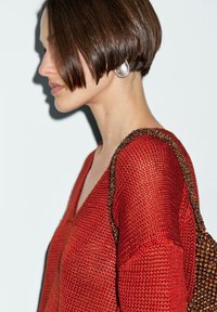 Top lavorato a maglia rosso con scollatura a V, caratterizzato da un motivo testurizzato e una tracolla perline marroni. Un orecchino ad anello argentato aggiunge un dettaglio metallico a contrasto.