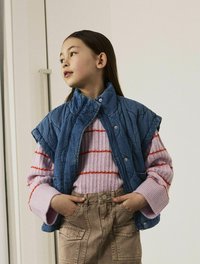 Džínová vesta s prešívaným vzorom, nosená cez ružový a červený pruhovaný sveter. Béžové nohavice majú vrecká a zapínanie na gombík.