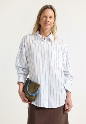 Kvinde med langt blondt hår iført en hvid skjorte med lyseblå striber, der holder en olivengrøn clutch med en blå rem.