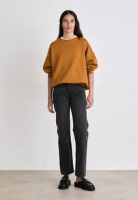 Suéter mostaza con un cuello blanco, jeans negros de pierna recta y mocasines negros. El modelo está de pie contra un fondo claro y liso.