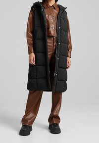 Gilet long noir rembourré avec une capuche, doté d'une fermeture éclair et de boutons-pression ; porté par-dessus une tenue en cuir marron et associé à des chaussures noires imposantes.