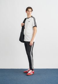 Biela krátka košeľa s čiernymi akcentmi, čierne teplákové nohavice s tromi bielymi pruhmi a červené tenisky s bielymi detailmi. Čierna ramenná taška.