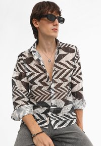 Camicia trasparente con colletto, caratterizzata da un motivo a zigzag nei colori nero, bianco e marrone, con chiusura a bottoni e maniche arrotolate.