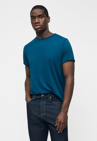 Άνδρας που φοράει ένα μπλουζάκι με κοντά μανίκια σε απόχρωση πετρόλ, κρυμμένο μέσα σε σκούρα μπλε τζιν, στέκεται με το ένα χέρι στην τσέπη, μπροστά από μια απλή, μονόχρωμη επιφάνεια.