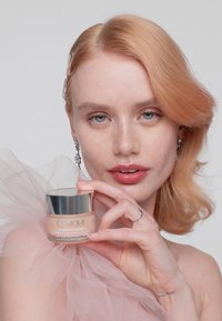 Pot de Clinique Moisture Surge avec un couvercle argenté, crème rose clair à l'intérieur. Le modèle tient le pot avec un tissu rose transparent et des bijoux subtils en arrière-plan.