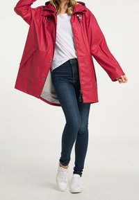 Impermeabile rosso con cappuccio, bottoni a pressione e due tasche laterali, indossato sopra una camicia bianca e jeans blu, abbinato a sneaker bianche.