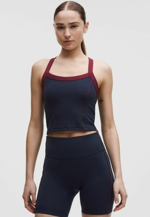 Młoda kobieta w dopasowanym sportowym topie w kolorze granatowym i bordowym oraz pasujących do niego wysokich spodenek rowerowych, stojąca na tle jednolitego tła.
