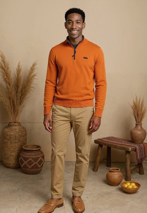 Pull à zip orange avec une accent noir, associé à un pantalon beige et des mocassins marron. L'arrière-plan présente des paniers tissés et des objets décoratifs.