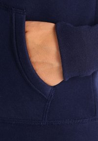 Mørkeblå hoodie med kengurulomme og ribbede manchetter. Stoffet ser blødt og tekstureret ud med synlige syninger.