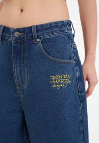 Denim jeans in dark blue, featuring a yellow embroidered logo on the front. Standard fit with five pockets and metal button closure.