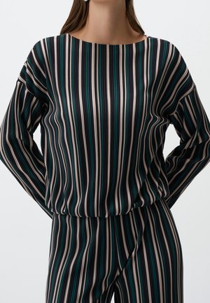 Femme portant un haut à manches longues et un pantalon à rayures verticales noires, blanches, bleu sarcelle et grises, debout les mains derrière le dos.